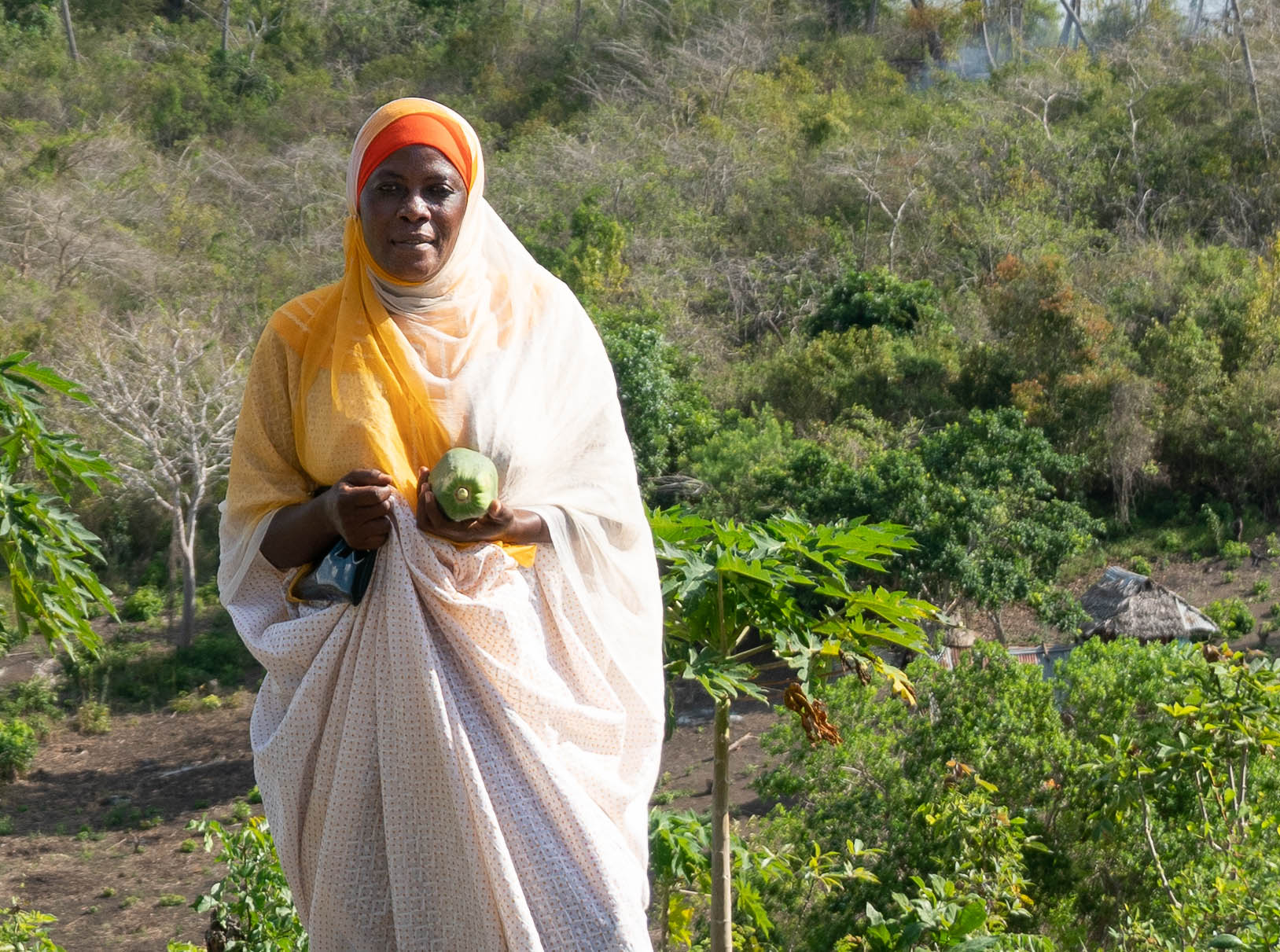 Woman in on a hillside in Tanzania holding her purse and a papaya