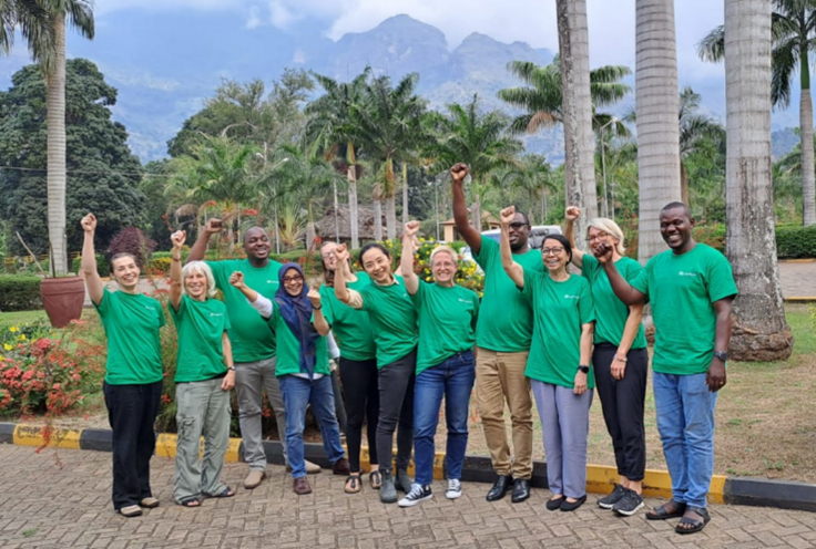 A diverse group of 11 Landesa staff members of the LPPG pose outside wearing matching green shirts and hold their arms up triumphantly while smiling.