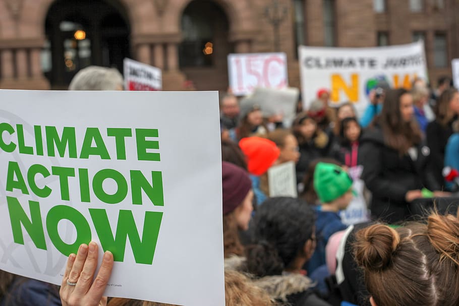 In the foreground, a hand holding a sign that says "CLIMATE ACTION NOW." In the background, a climate strike in Toronto.